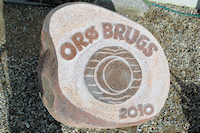 Ved udgravning af
grunden til den nye Brugs p� Or� blev fundet en r�d Halmstad-granitsten. Stenen
er mange millioner �r gammel, men vandringen mod Or� startede m�ske
for 100.000 �r siden, da den f�rste istid l� tungt over Skandinavien og begyndte
at ase og mase med denne sten oppe fra Hallands�sen i Sverige ned
mod Or�. Stenens v�gt er ca. 1500 kg. Jeg t�nkte �Hvorfor ikke en inskription p� stenen
foran den nye Brugs?� En stenskulptur er normalt noget, der st�r ved en
offentlig bygning, men hvorfor ikke foran en Brugs i �Udkantsdanmark�? S�
gik jeg i gang med at lave skitser. Symbolet
har jeg f�et inspiration til p� Stenhuggerskolen, det viser solen som det
livgivende, m�nen som det cykliske og det tilbagevendende, og vandet som
det vi alle kommer fra.
Teksten p� stenen er et udtryk for �boernes selvst�ndighed. Det er
Or�s Brugs, ikke Coop Danmarks.